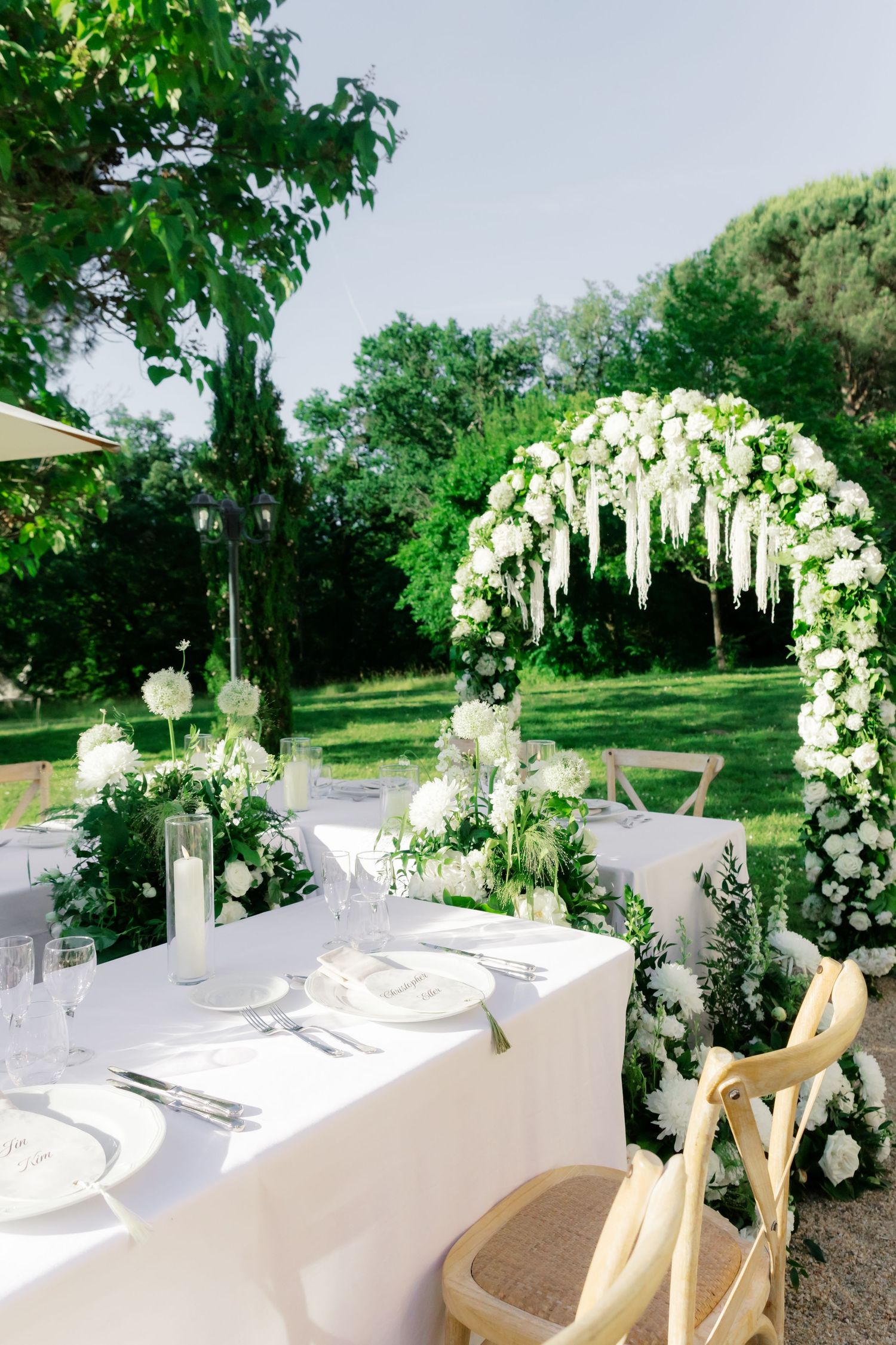 Elegant outdoor wedding reception with white floral arch, draped blooms, and a beautifully set dining table in a lush green garden.