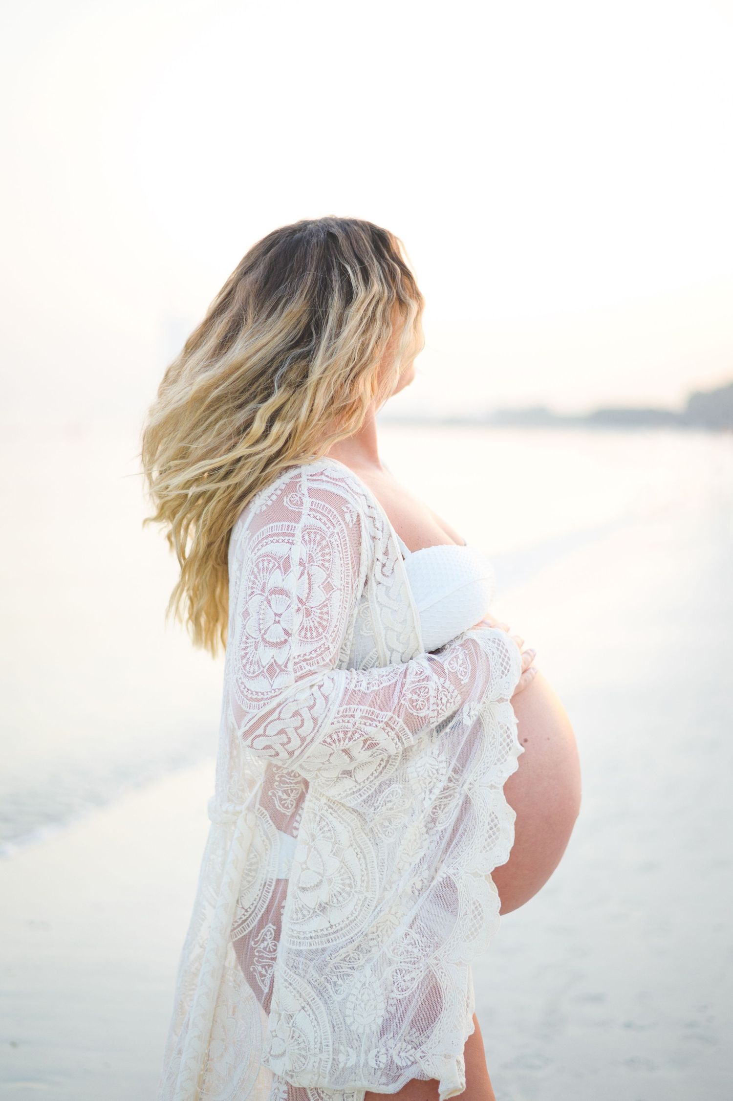 Pregnant woman in white lace robe cradling her baby bump on a serene beach at golden hour.