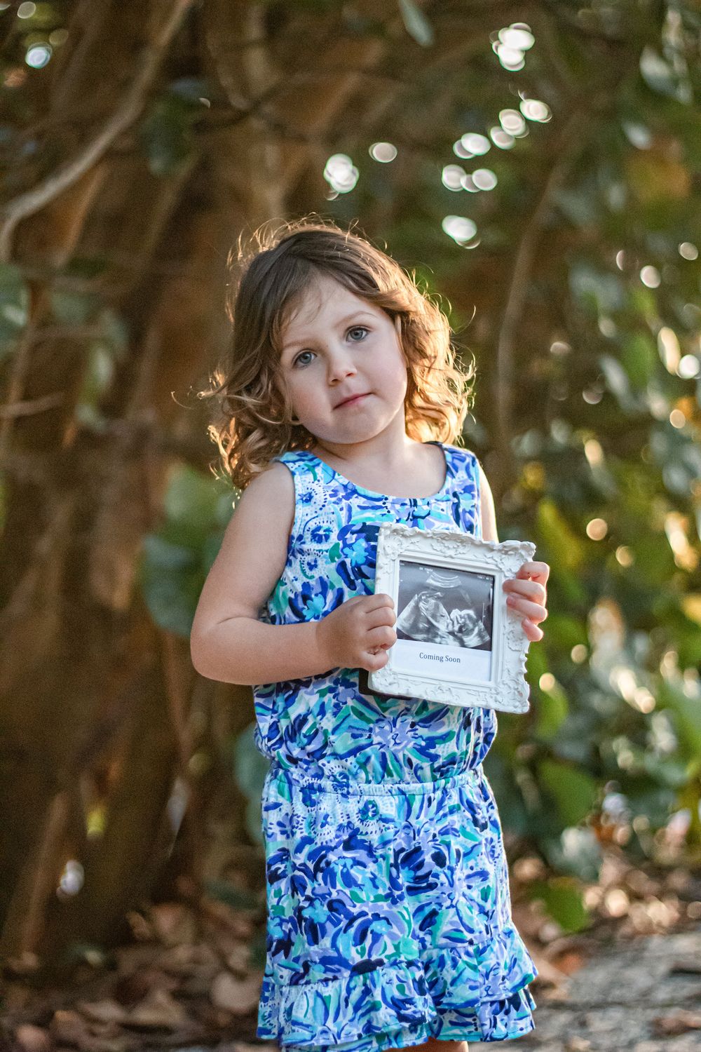 Young girl in blue floral romper holding a framed ultrasound photo outdoors among trees Blowing rocks beach Jupiter.