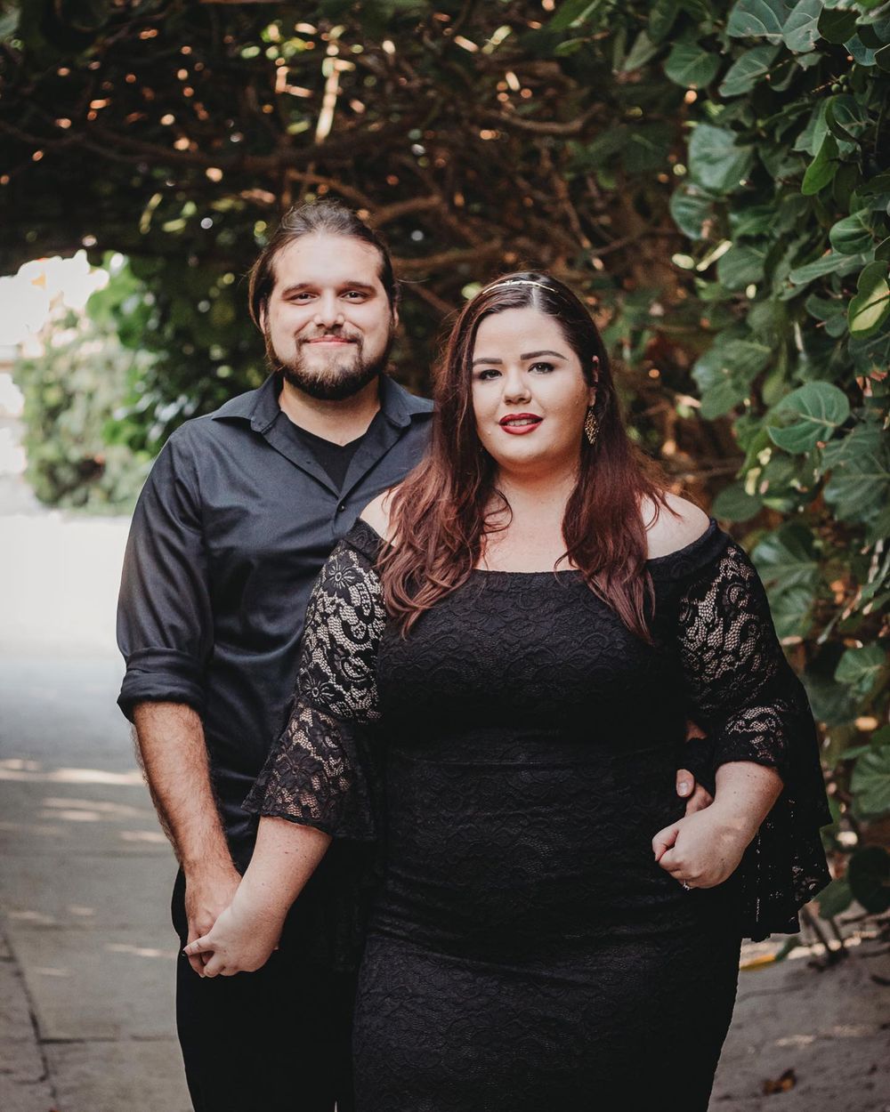 A couple in matching black outfits holds hands while posing under a lush green archway on a garden path at Blowing rocks beach Jupiter.