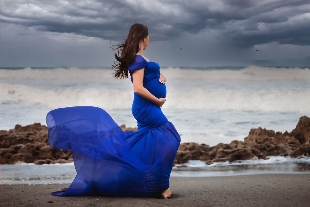 Pregnant woman in flowing blue gown poses on rocky beach with stormy sky and crashing waves behind her on a beach in Boynton.