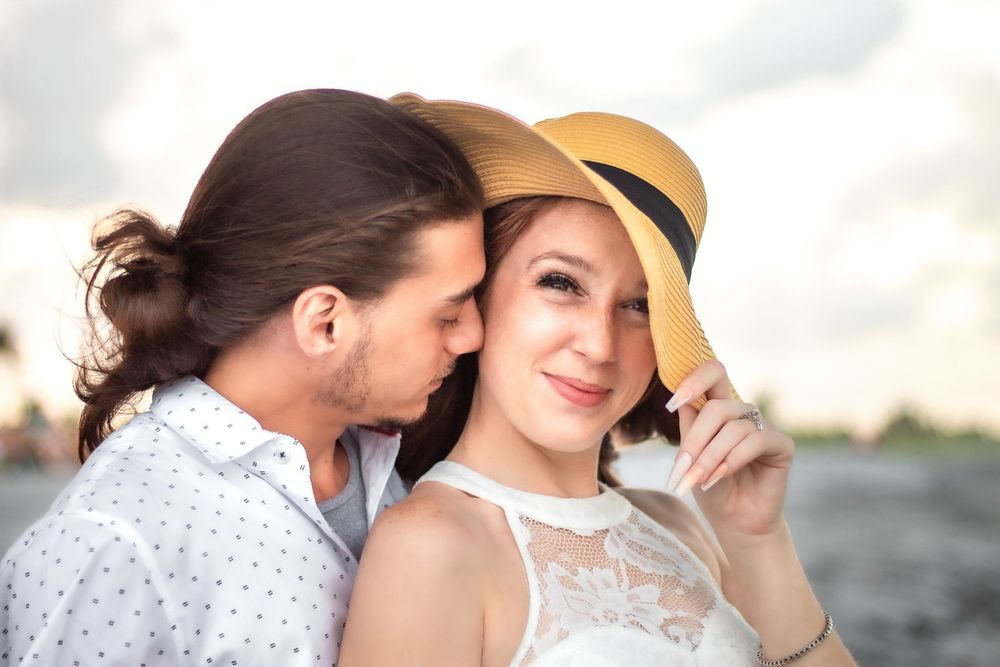 Romantic couple outdoors, man whispering to smiling woman in straw hat near waterfront in palm beach.
