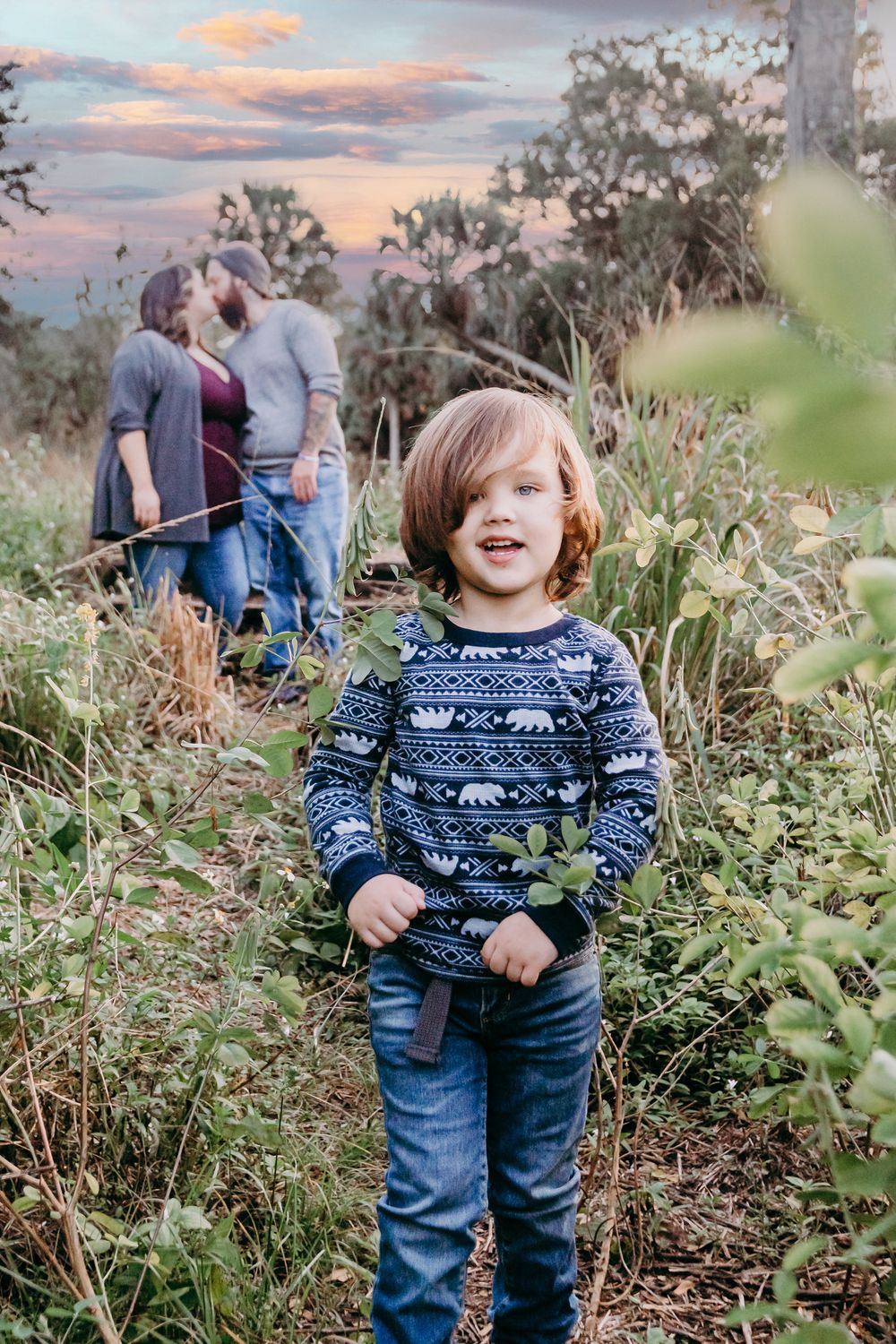 Young boy in blue patterned sweater and jeans smiles outdoors at sunset, couple kissing in background at riverbend park Jupiter