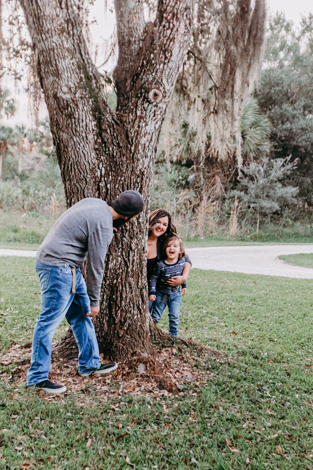 Family of three playing peek-a-boo around a large tree in a park, with a laughing toddler and smiling parents at riverbend park Jupiter.