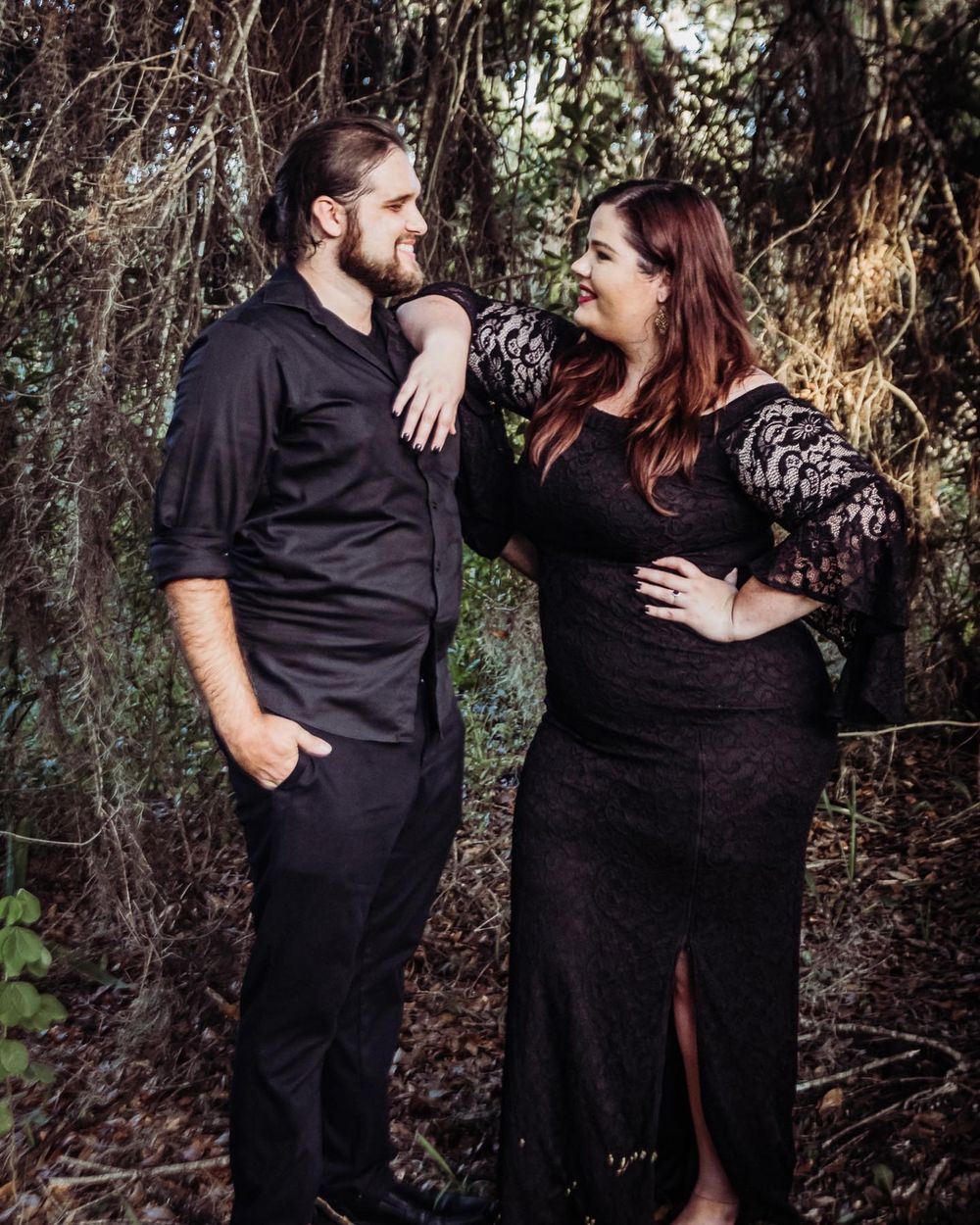 Couple in matching black outfits posing romantically in a wooded outdoor setting at riverbend park.