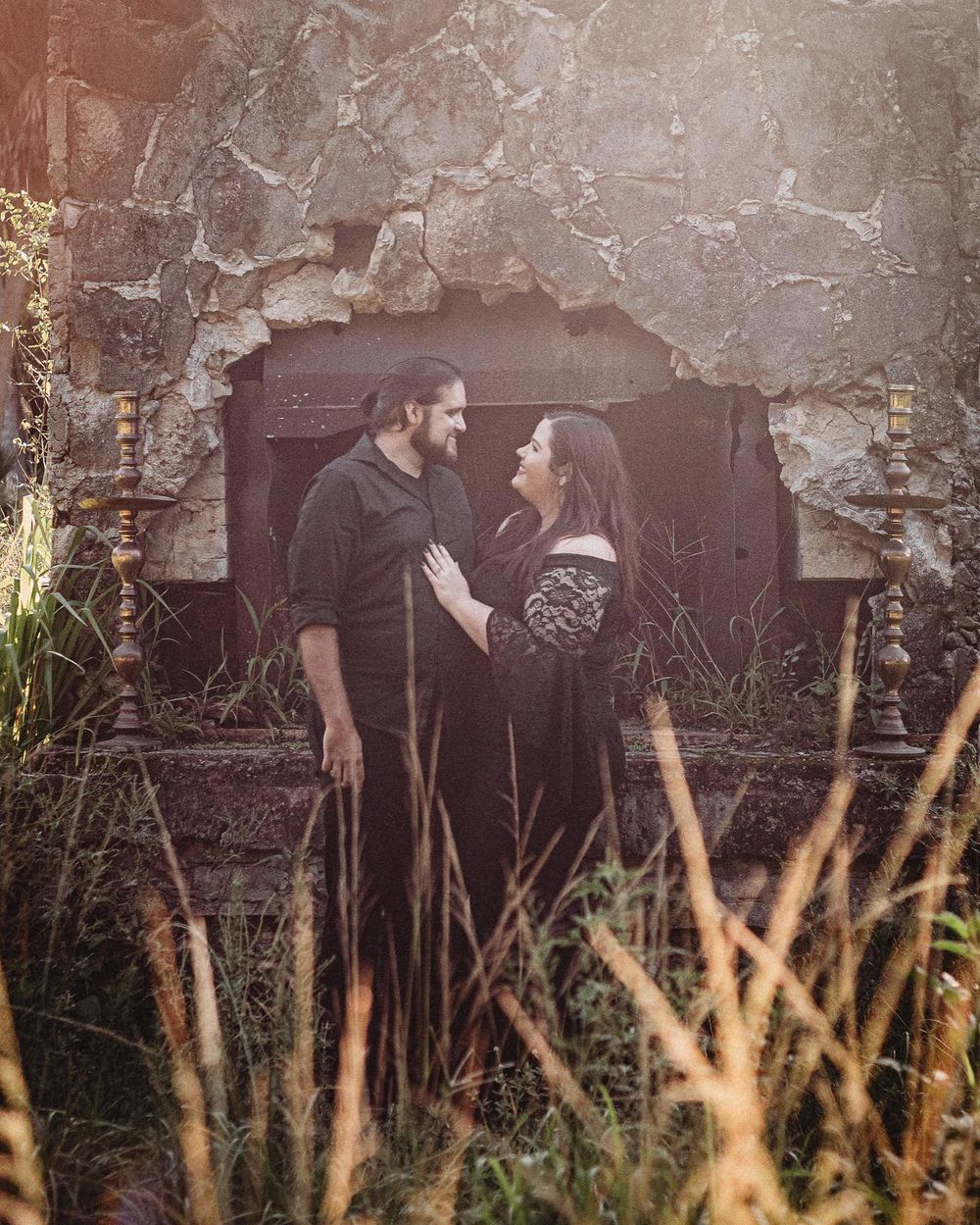 Couple in black attire gazing at each other before rustic stone ruins surrounded by golden wild grass at riverbend park.