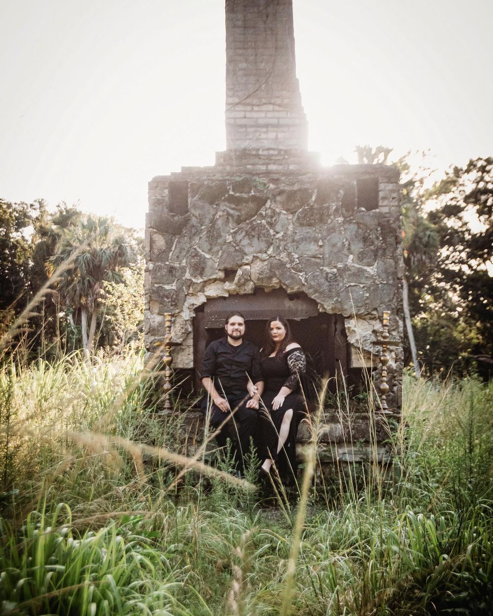Couple sitting together in front of a rustic stone fireplace surrounded by lush greenery in a scenic outdoor setting at riverbend park.