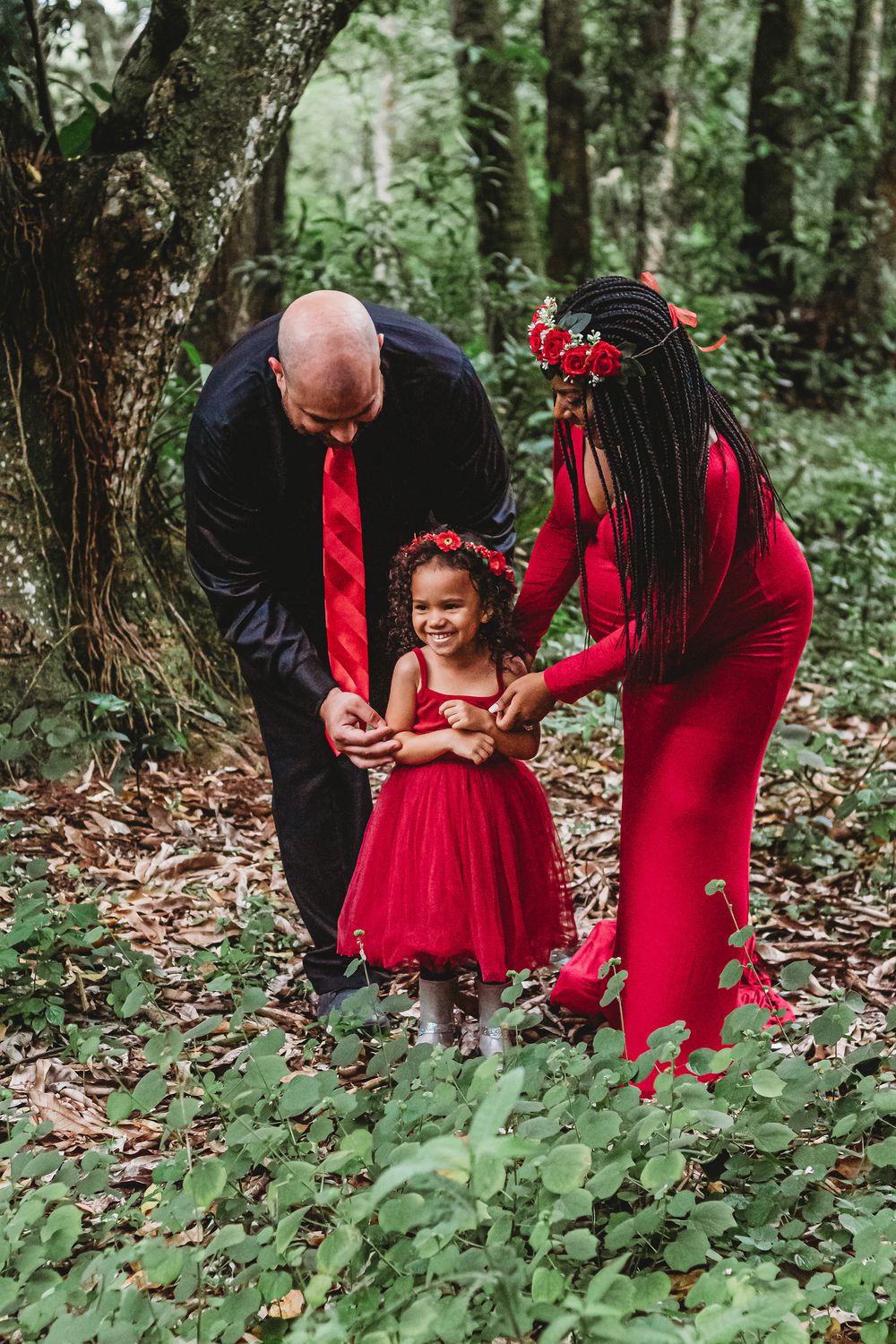 A family in matching red outfits poses in a lush forest, with parents holding their smiling young daughter's hands at riverbend park Jupiter