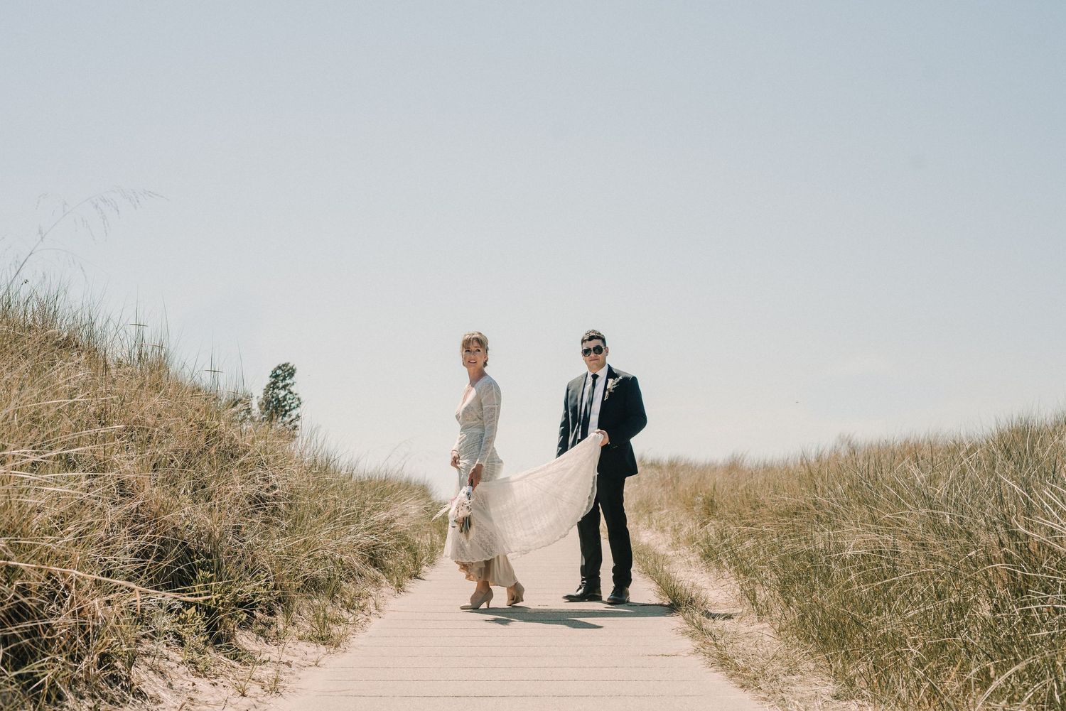 A couple in wedding attire walks along a coastal boardwalk pathway surrounded by tall dry grass on a sunny day.