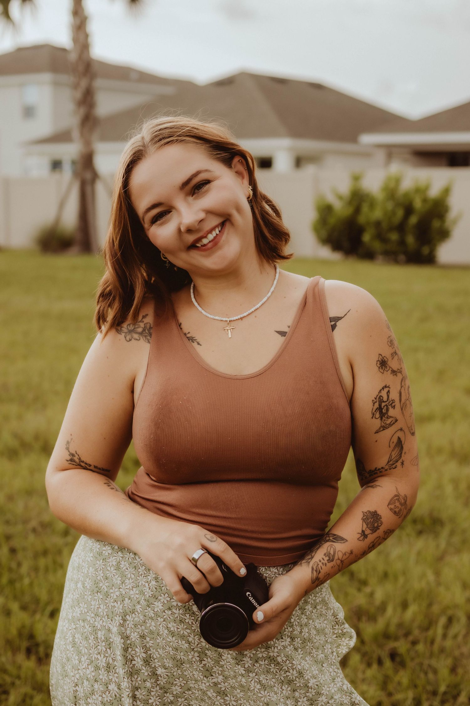 Smiling young woman with tattoos wearing a rust tank top and floral skirt, holding a camera lens while sitting on green grass.