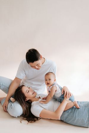 Happy young Asian family with baby relaxing on floor, parents in white tees and jeans in bright studio setting.