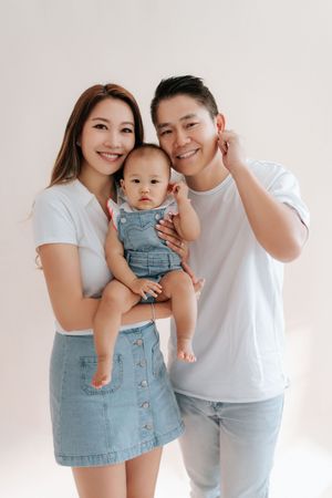 Asian family portrait with smiling parents holding their baby in denim outfits against a white background.