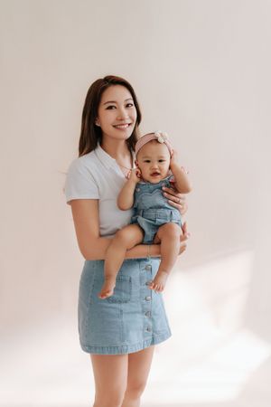 Smiling Asian mother in white top and denim skirt holding her baby girl in denim romper with pink bow headband.