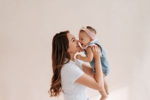 A loving mother holds her smiling baby girl up close for a kiss against a soft neutral background.