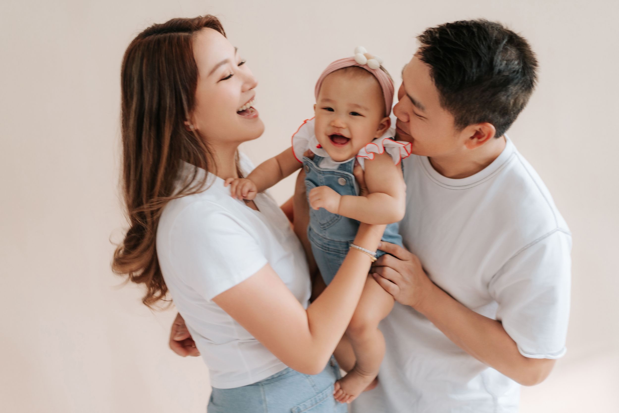 Happy Asian family with smiling parents holding their laughing baby girl in a bright studio setting.