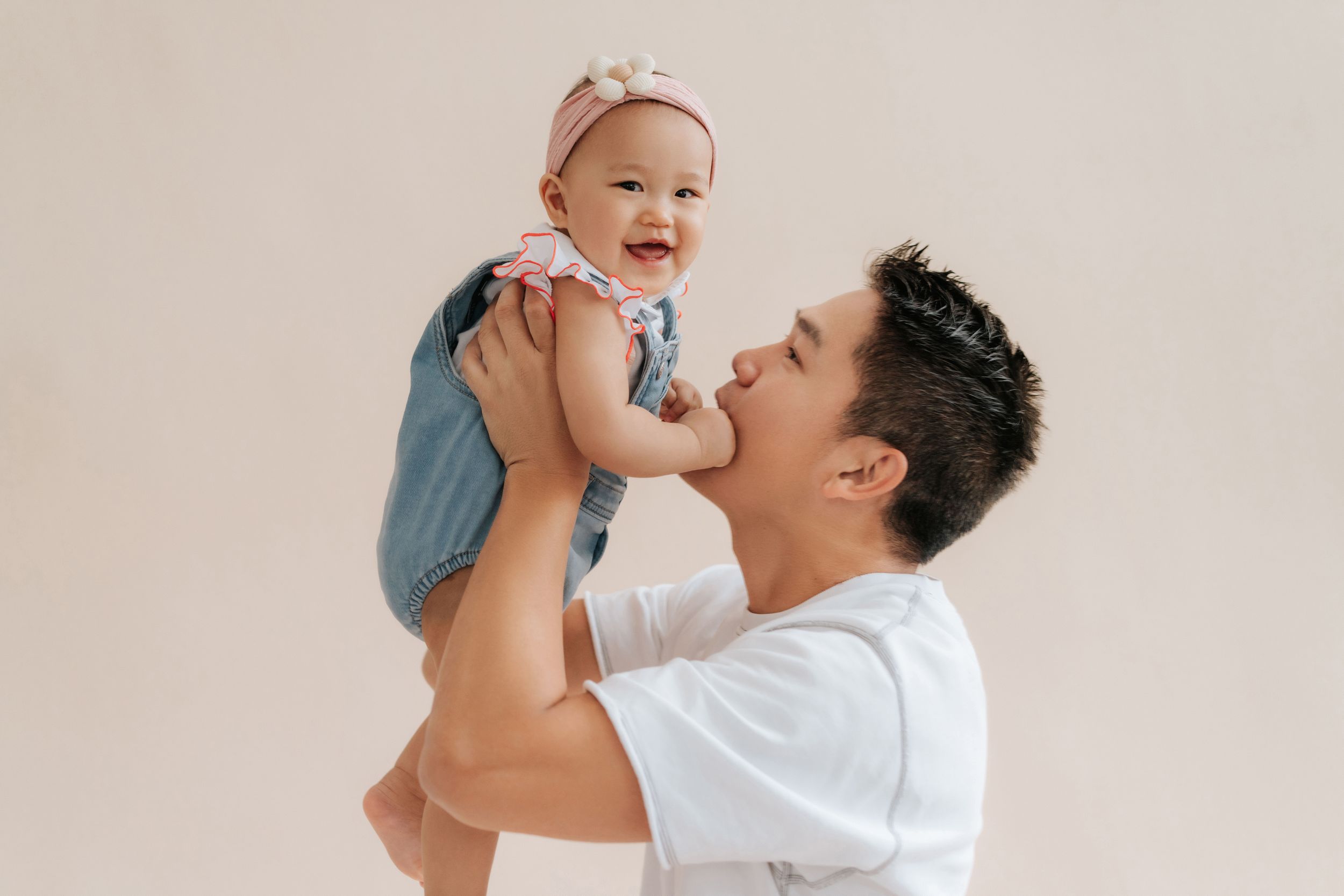 A smiling man in a white shirt lifts a laughing baby girl wearing a pink bow and denim outfit against a beige background.