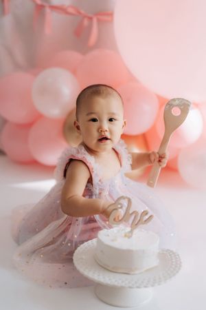 Baby girl in pink tutu dress celebrates first birthday with white cake and wooden spoon, surrounded by pink balloons.