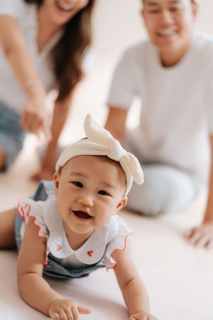 A happy baby girl in denim overalls and white bow headband crawls toward camera as smiling parents watch from behind.