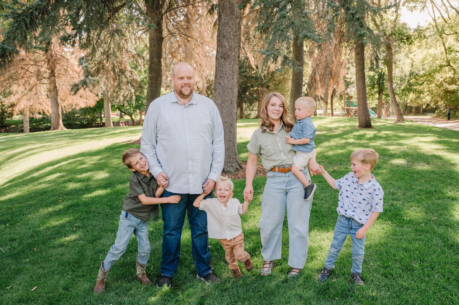 A family poses together on a sunny day in a grassy park with tall trees in the background.