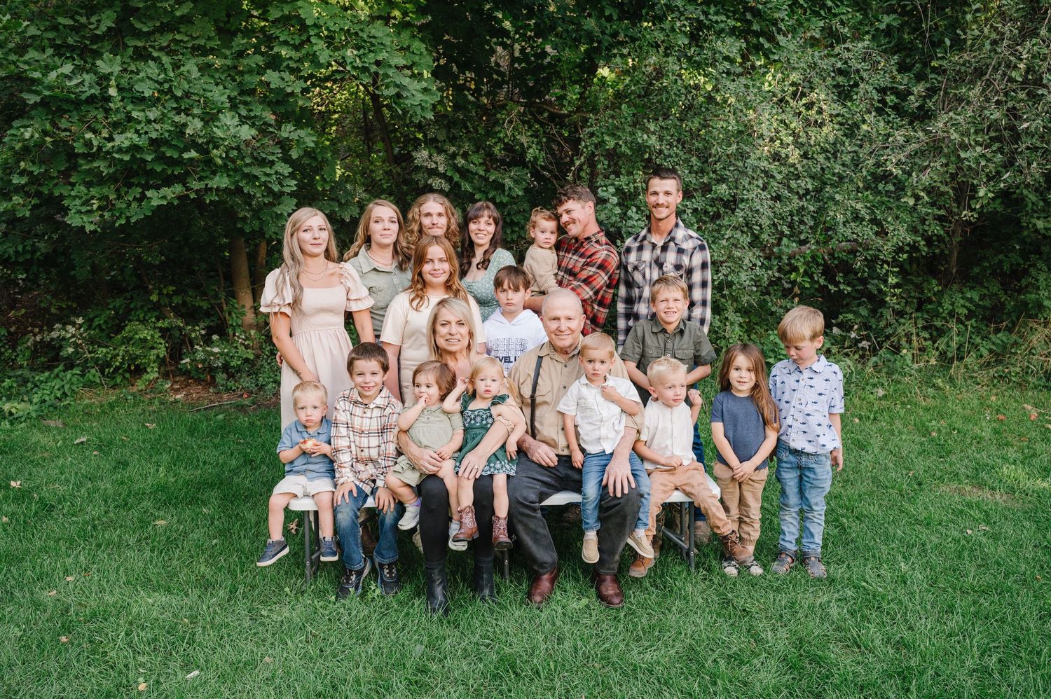 A group of children and adults posed together outdoors on a sunny day in front of green foliage.