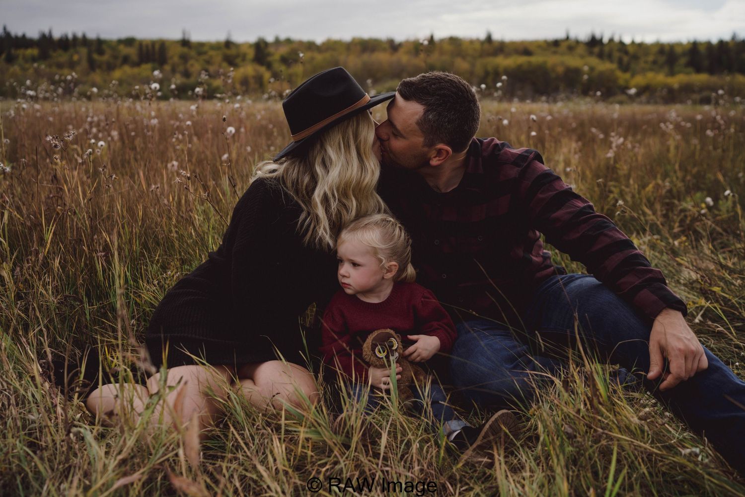 A couple sits in a golden field, sharing a kiss over their toddler in a cozy autumn family portrait.