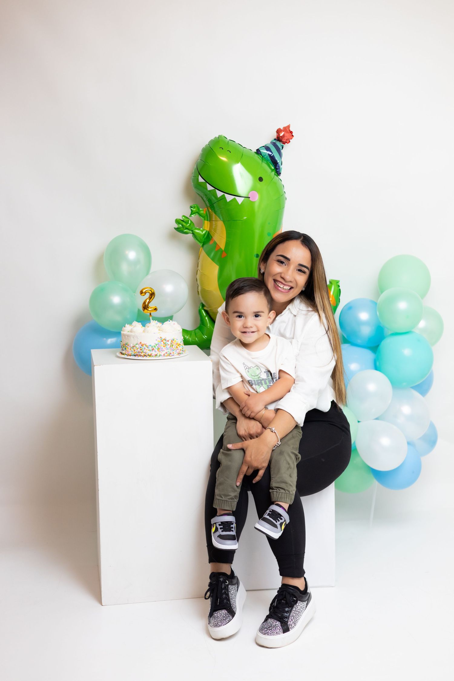A mother and toddler celebrate a 2nd birthday with a dinosaur balloon, cake, and blue and green balloon decorations.