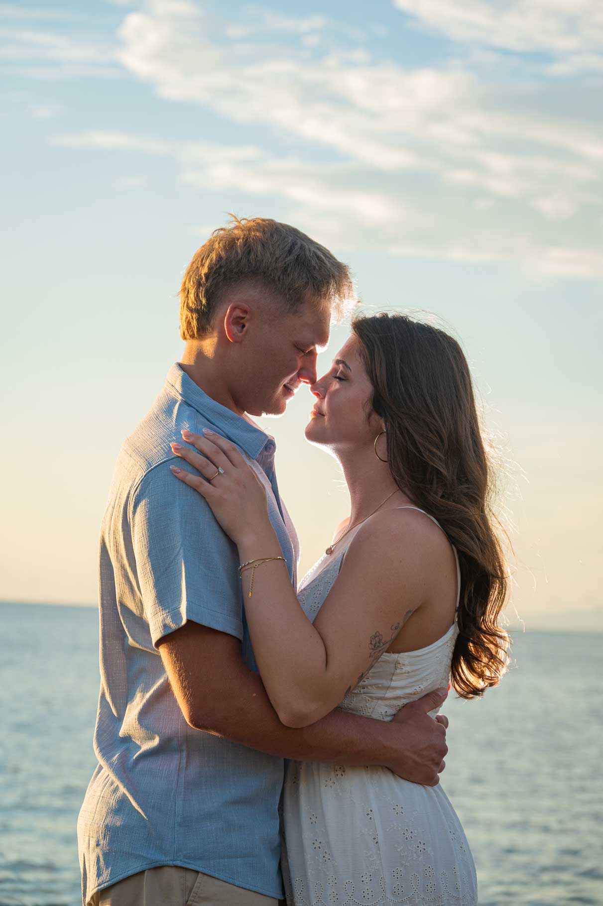 A romantic couple embraces and nearly kisses at sunset by the ocean, dressed in casual white and blue attire.