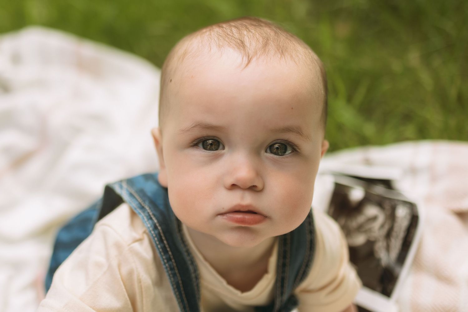 Adorable baby in denim overalls lying on a blanket in green grass, looking up with curious eyes.