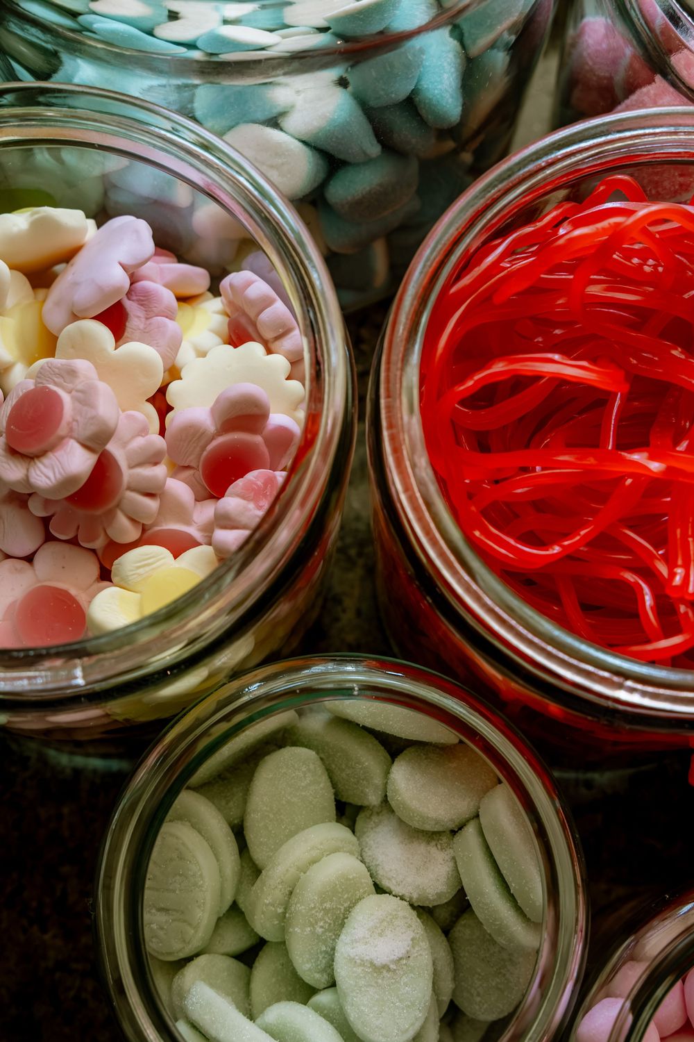 Glass jars filled with colorful assorted candies including red licorice, mint discs, and flower-shaped sweets on a dark surface.