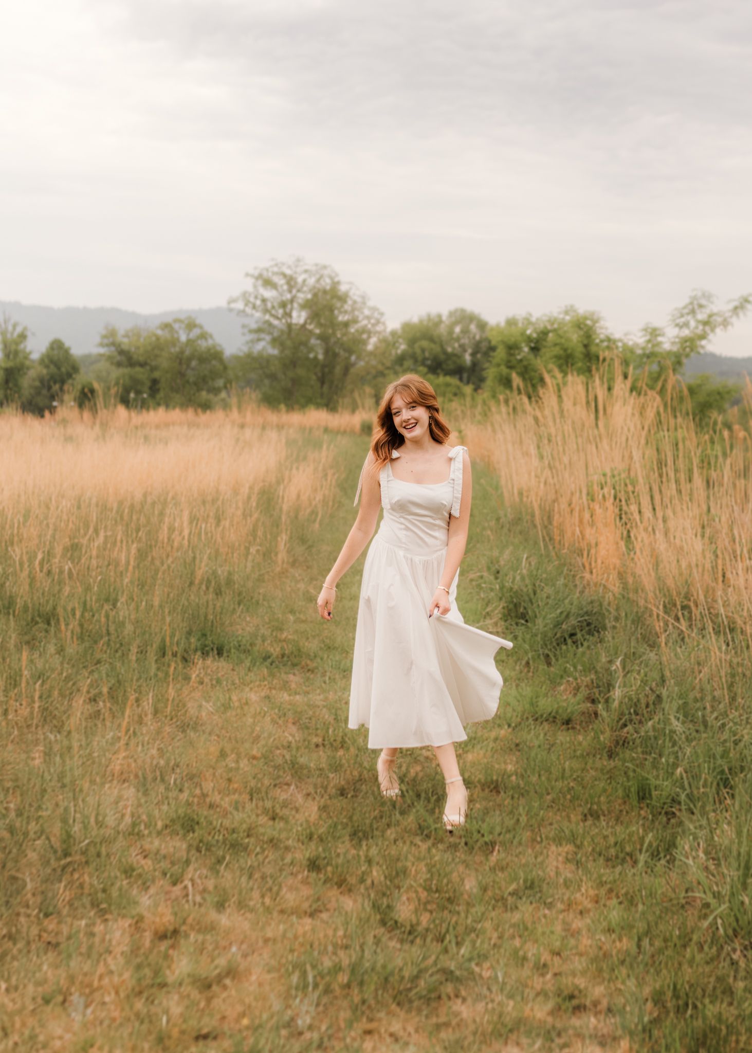 Woman in white midi dress walking through golden meadow with mountains and trees in background.
