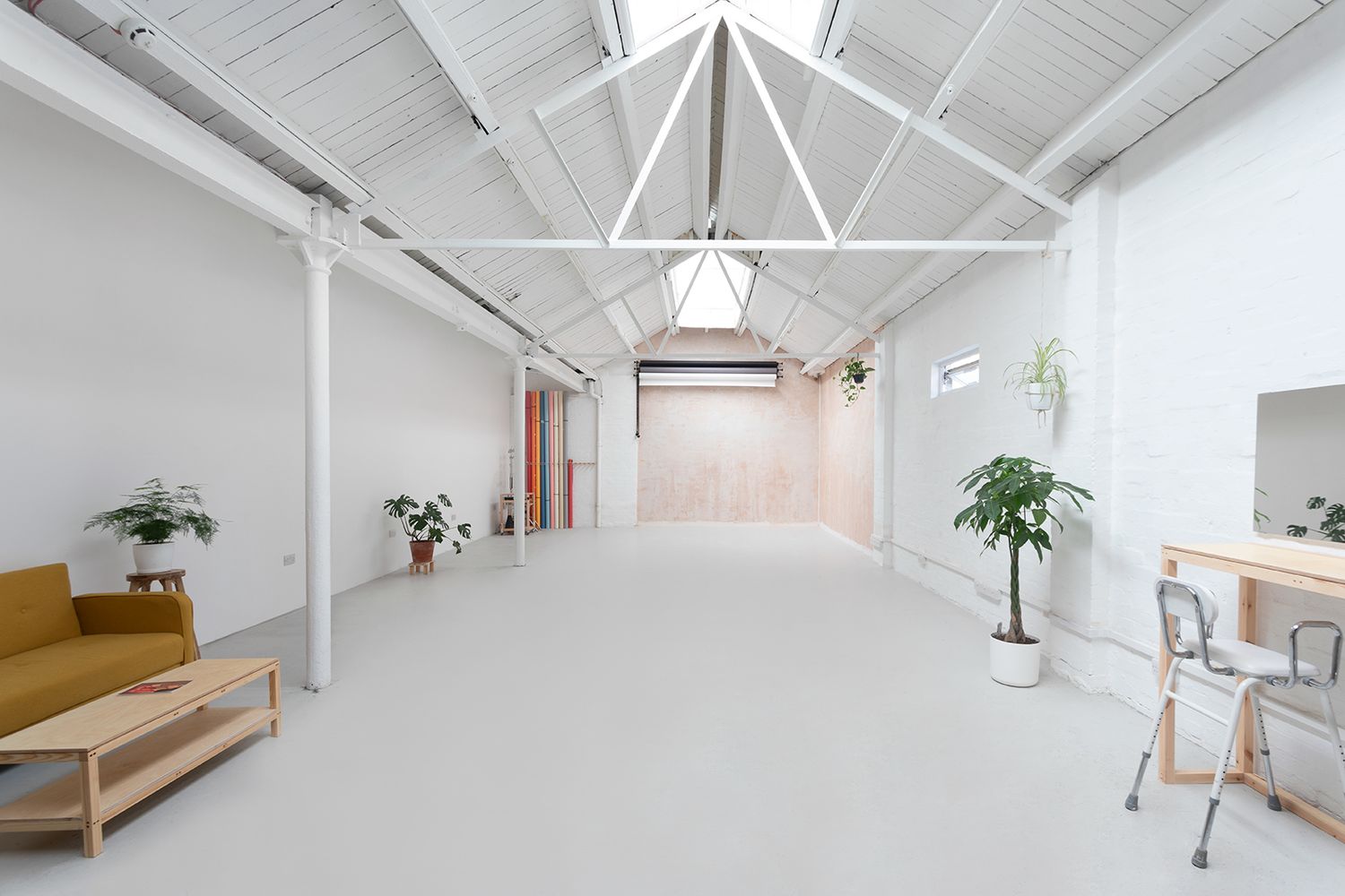 Bright white studio space with high vaulted ceiling, yellow sofa, wooden furniture, and lush green plants.