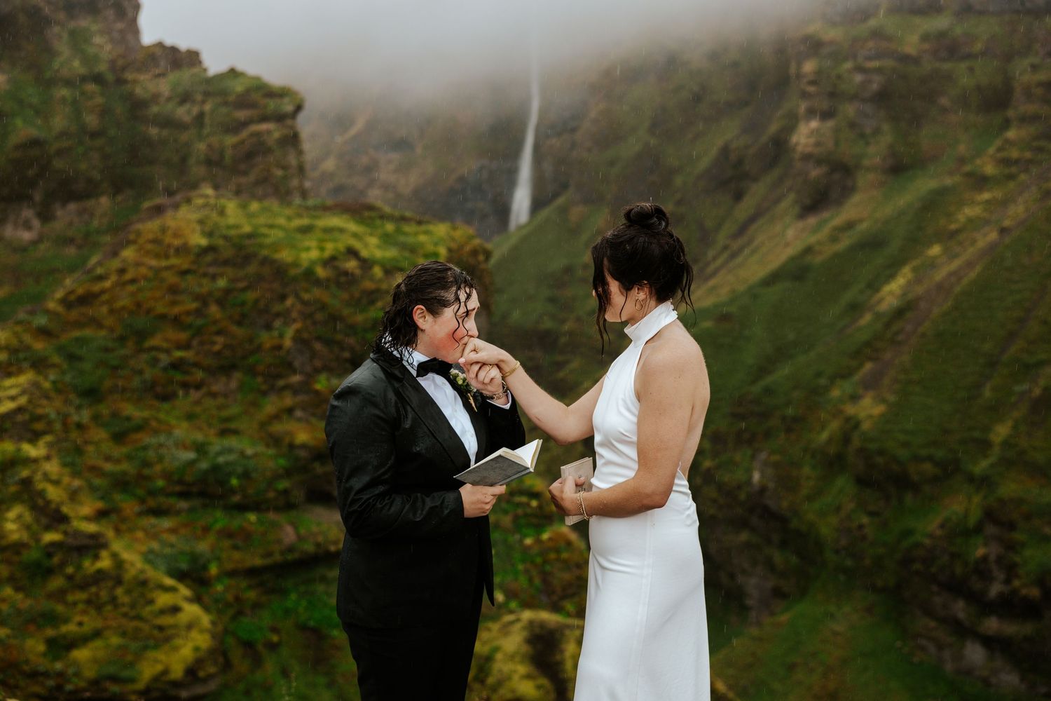 bride reading vows and kissing the hand of her bride while overlooking Mulagljufur Canyon and waterfall in Iceland during their elopement