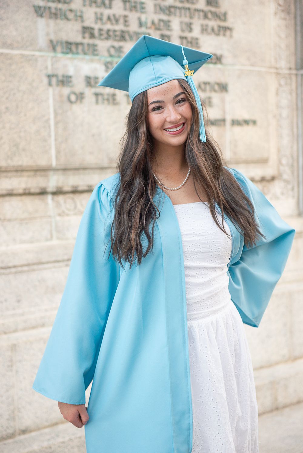 Smiling young woman in light blue graduation cap and gown posing outdoors in front of a stone monument.