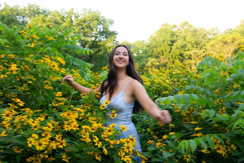 A young woman in a blue dress smiles joyfully among vibrant yellow wildflowers and lush green foliage outdoors.