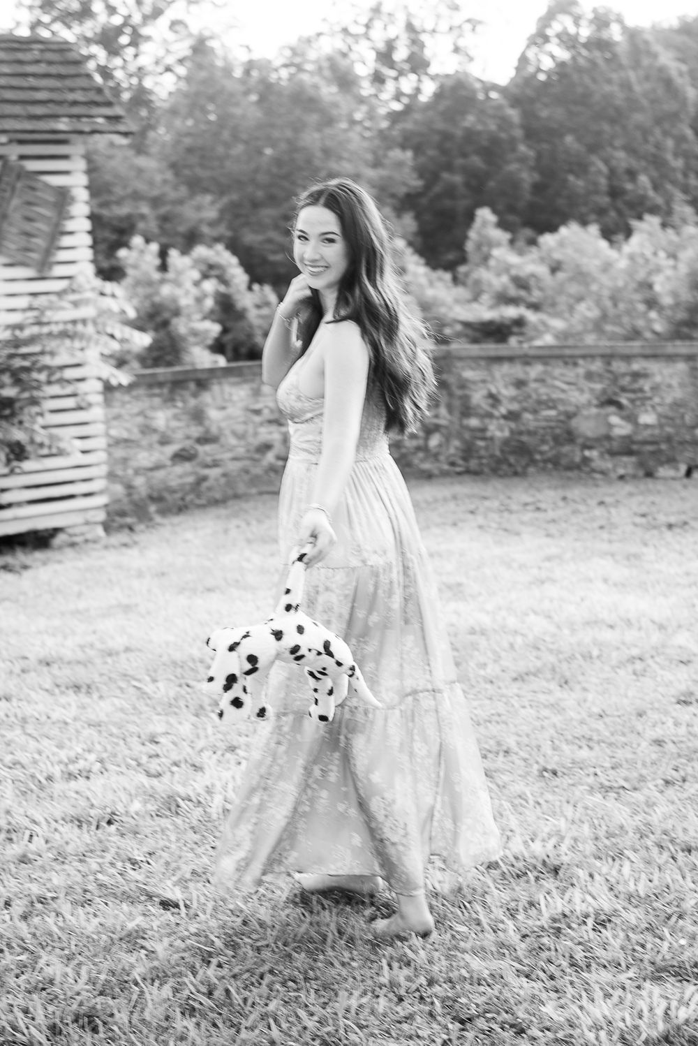 Black and white photo of a woman in a long flowy dress walking on grass, holding a Dalmatian puppy outdoors.