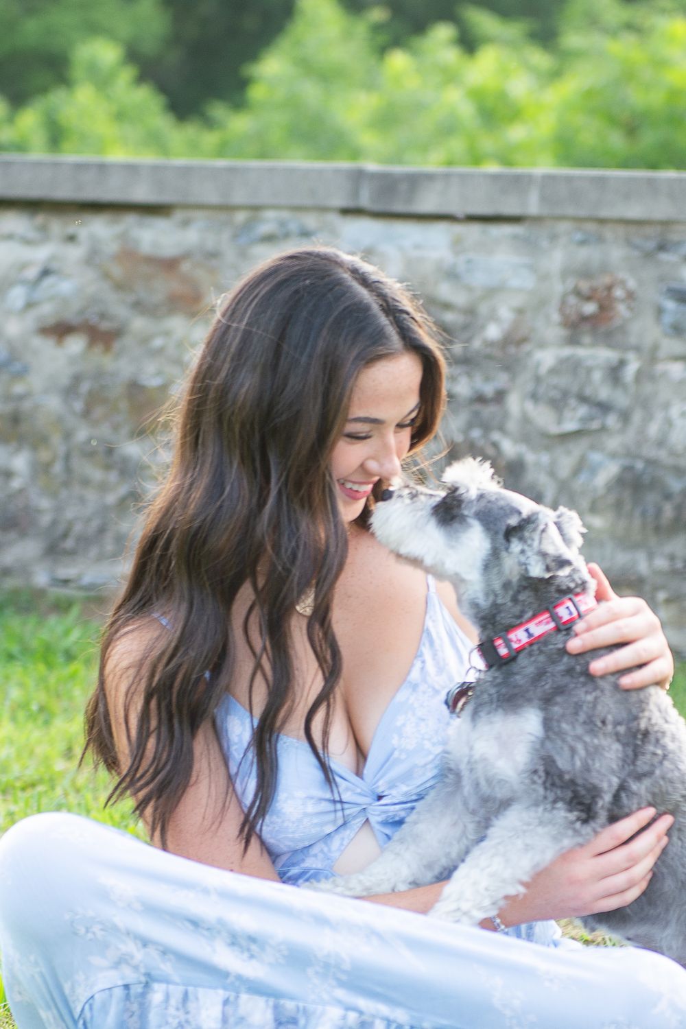 A smiling young woman with long dark hair sits on grass holding a gray schnauzer dog wearing a pink collar.