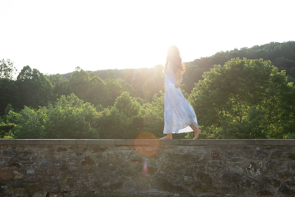 Woman in white dress standing on stone wall at sunset with lush green trees in background.