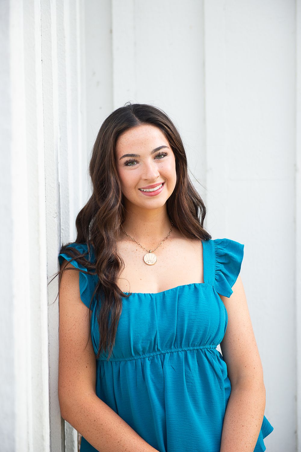 Young woman in teal ruffled dress and gold necklace smiling while leaning against a white column.