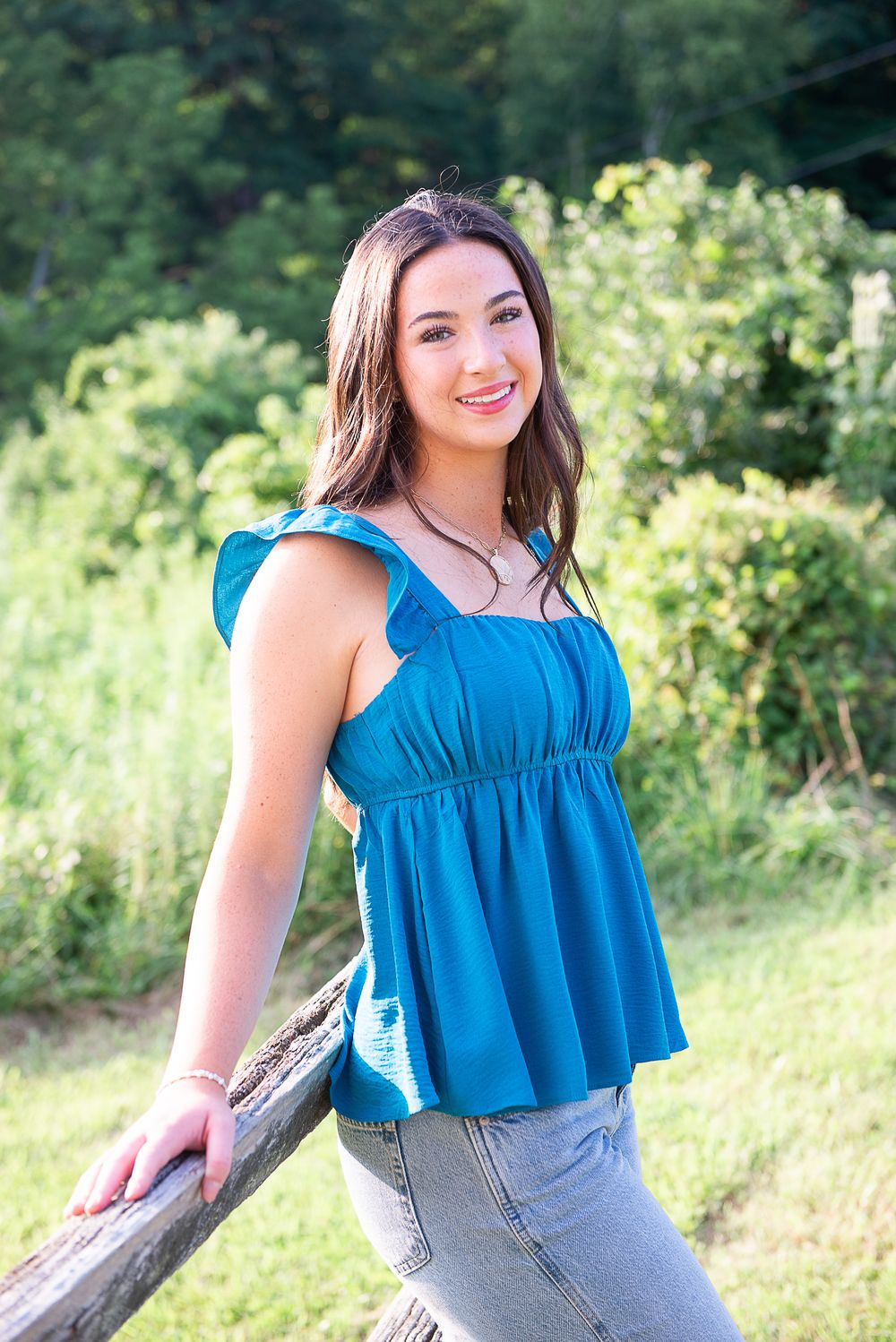 Young woman in teal ruffled top and jeans leaning on wooden fence outdoors.