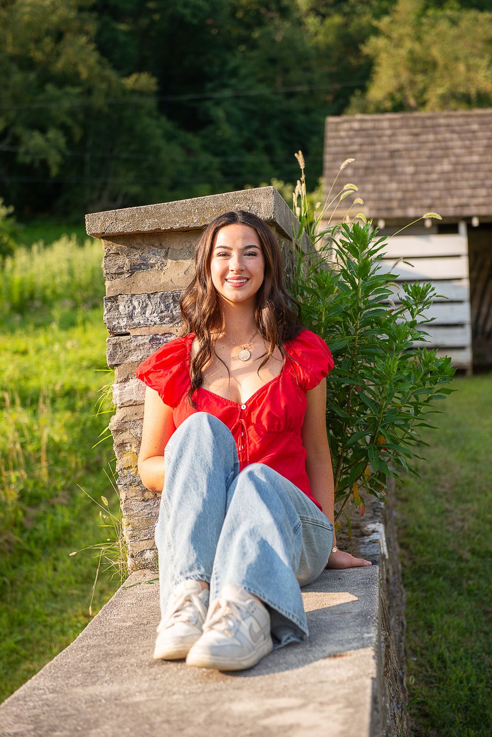 Young woman in red top and jeans sitting on stone wall outdoors, smiling, with green trees and rustic cabin background.