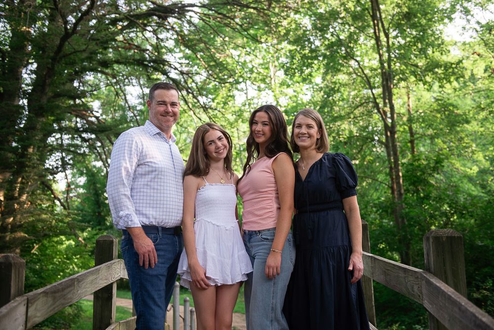 Family of four smiling together on a wooden bridge surrounded by lush green trees in a park setting.