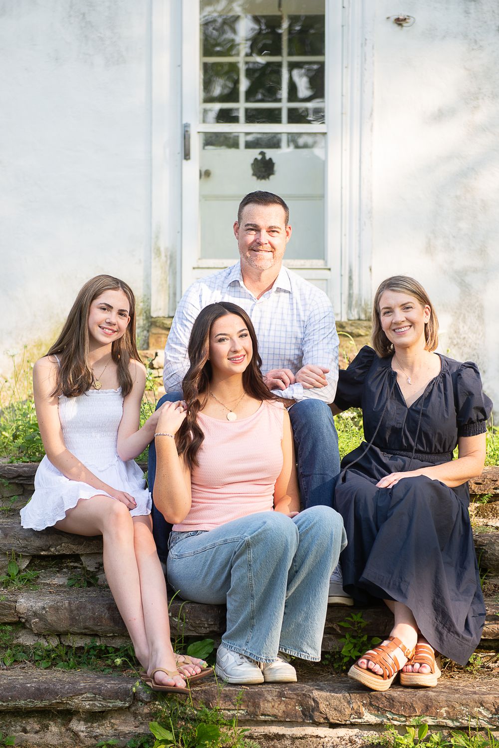 Family of four smiling together on stone steps in front of a white building in a candid outdoor portrait.