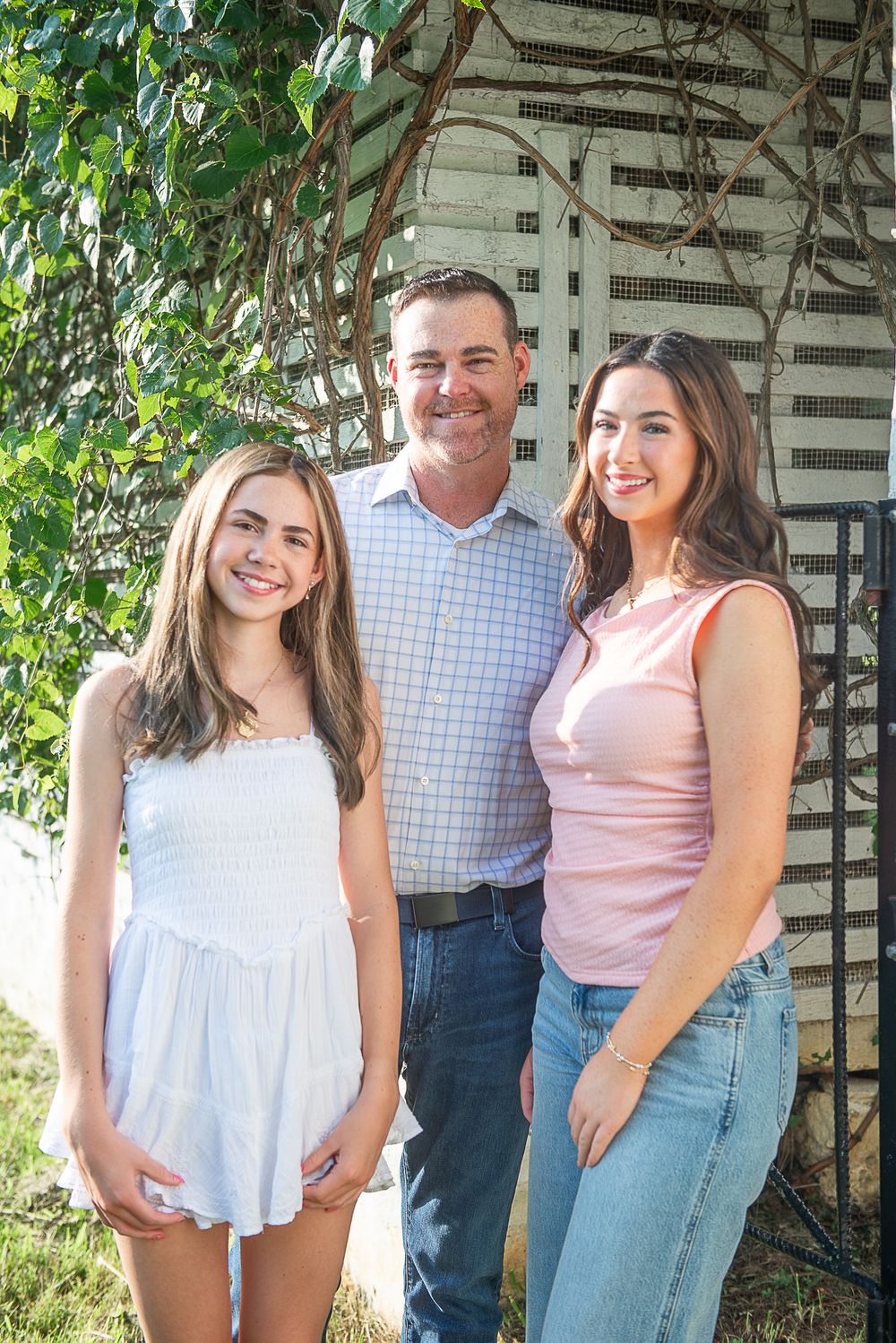 A man and two young women smile together outdoors near a white lattice structure with green vines.