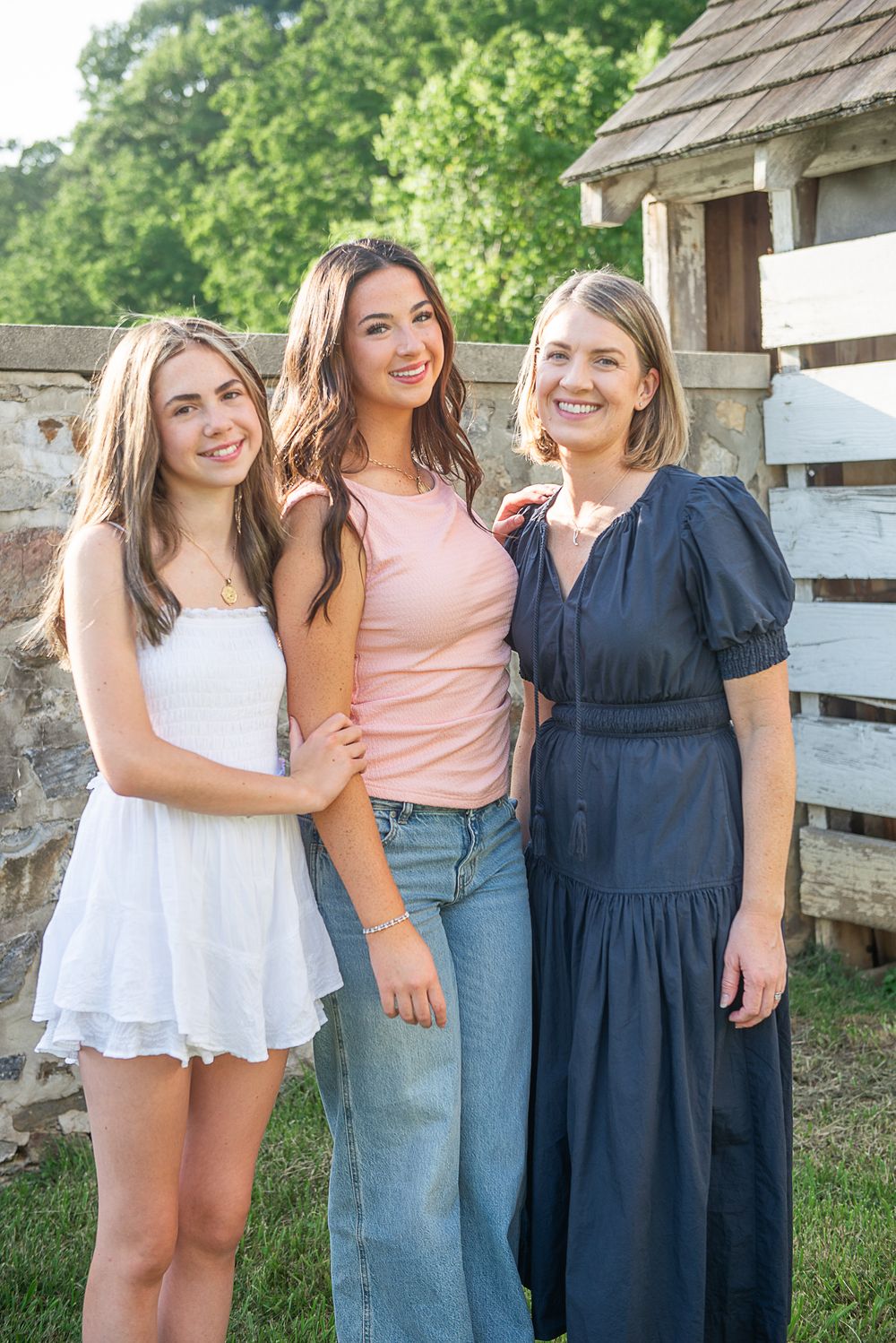Three smiling women pose together outdoors near a rustic wooden fence on a sunny day.
