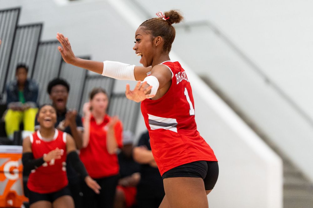 A volleyball player in red uniform celebrates a point with teammates cheering on the bench.