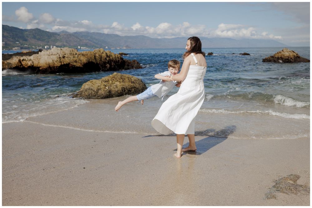 Family Vacation Photos at Conchas Chinas Beach in Puerto Vallarta with a mother in a white dress playfully twirling her son beside.