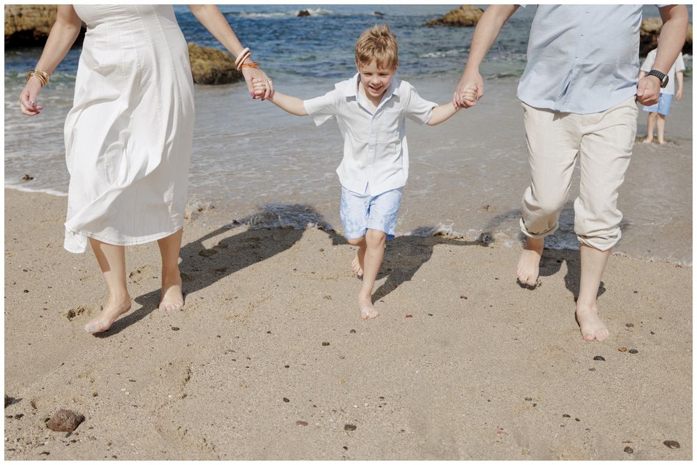Family Photoshoot at Conchas Chinas Beach in Puerto Vallarta with parents joyfully swinging their son barefoot along the sunny shoreline.