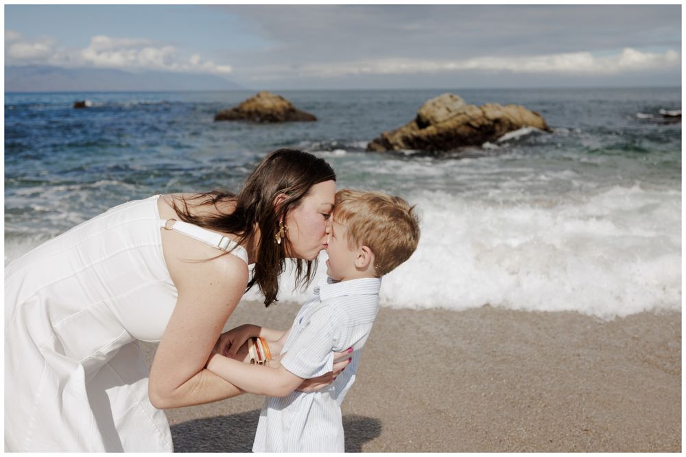 Family Beach Photos at Conchas Chinas Beach in Puerto Vallarta capturing a mother tenderly kissing her son beside the blue ocean waves.
