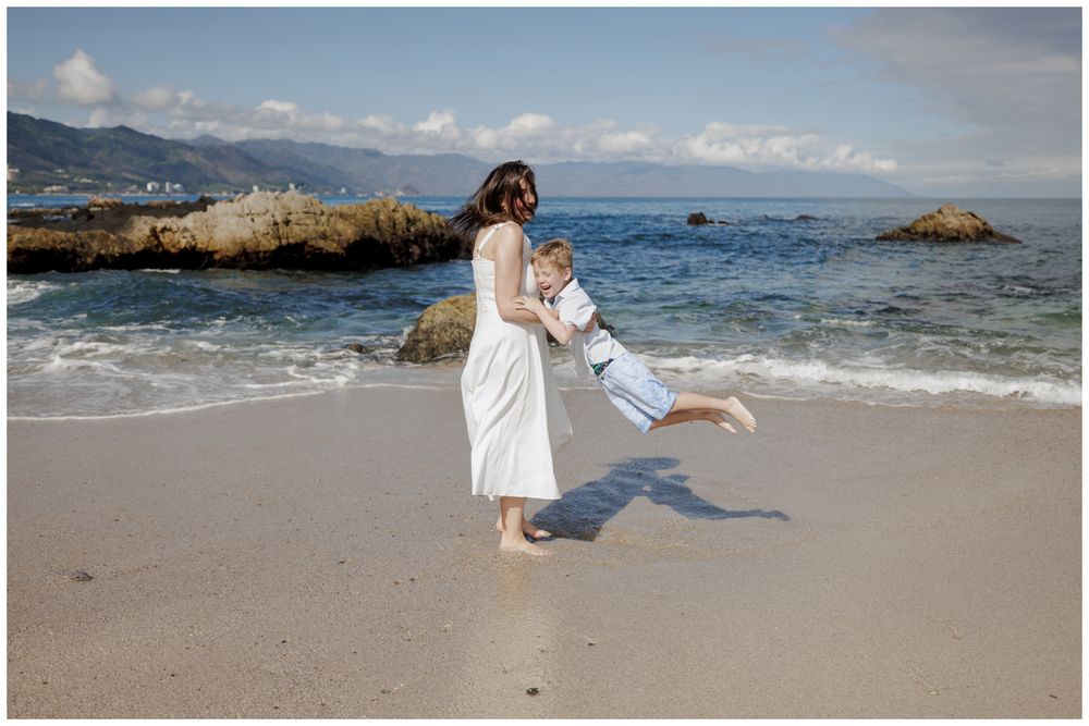Beach Photo Session at Conchas Chinas Beach in Puerto Vallarta with a mother playfully swinging her son along the sandy shoreline.