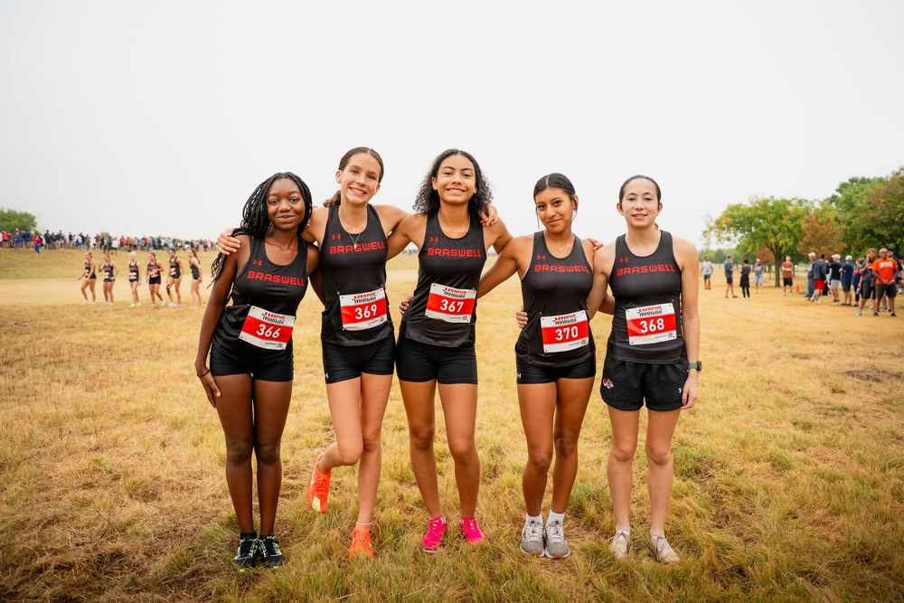 Five female cross country runners pose together wearing race bibs at outdoor meet.
